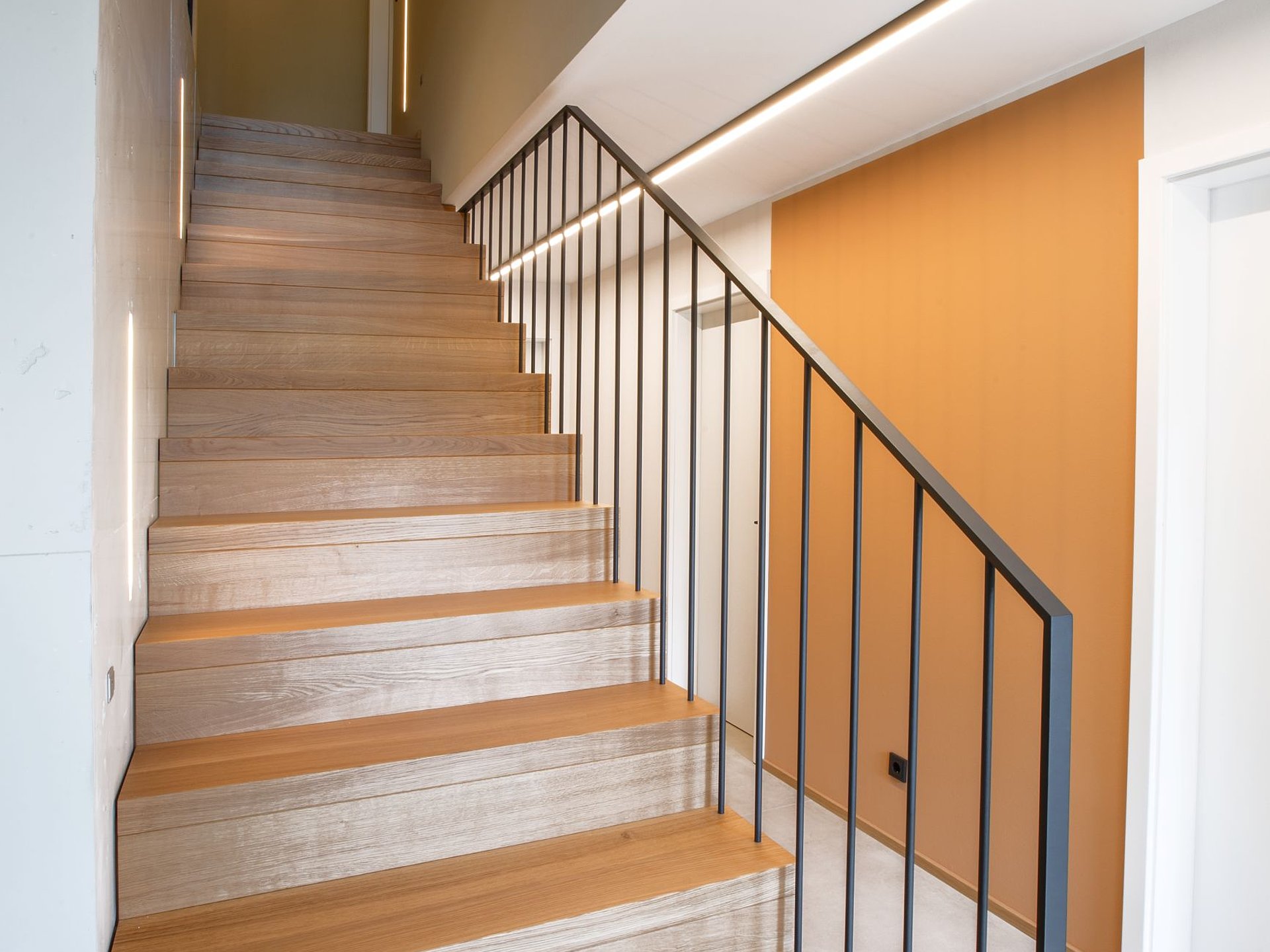Holztreppe mit filigranem Staketengeländer Holztreppe mit geschlossenen Setzstufen und filigranem, anthrazitfarbenem Staketengeländer. Eine lineare Beleuchtung begleitet den Treppenlauf.