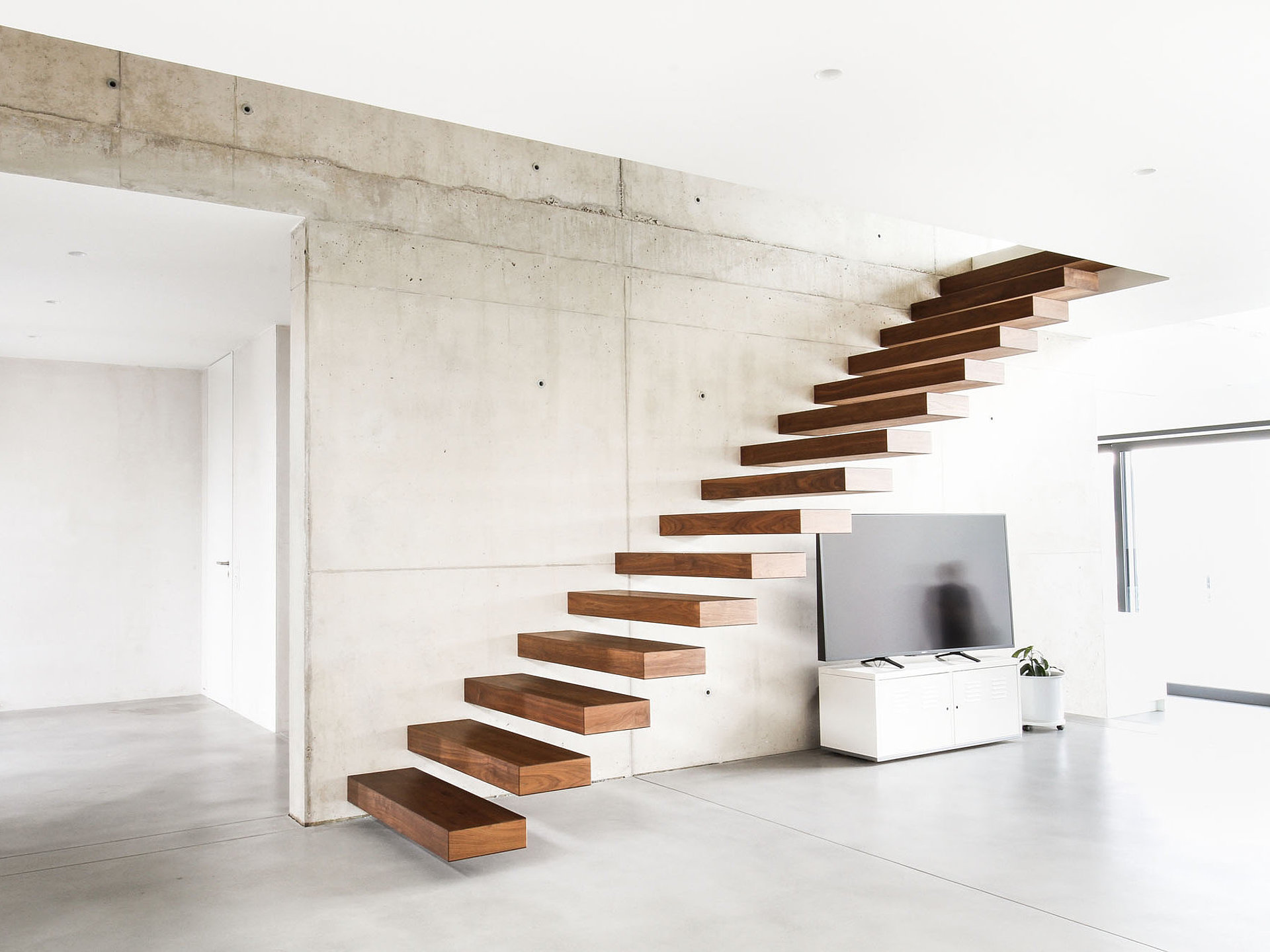 Kragarmtreppe n Nussbaum an Sichtbetonwand Minimalistische Kragarmtreppe mit dunklen Holzstufen an einer Sichtbetonwand. Die schwebende Konstruktion unterstreicht das moderne Wohnkonzept.