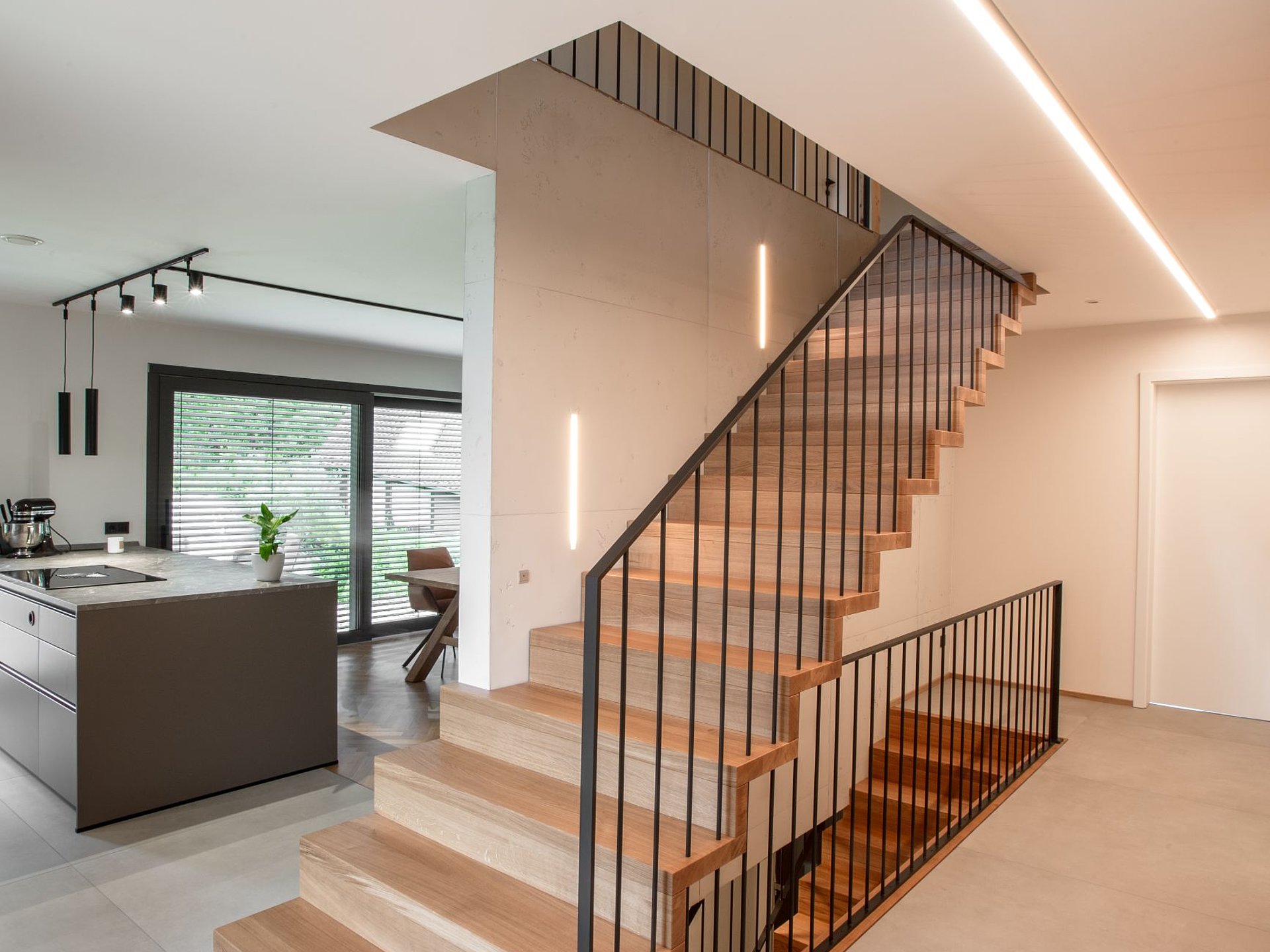 Moderne Faltwerktreppe aus Eichenholz mit geschlossen wirkenden Stufen und Setzstufen sowie filigranem, dunklem Staketengeländer. Sie verbindet zwei Wohnebenen in einem offenen Raumkonzept.